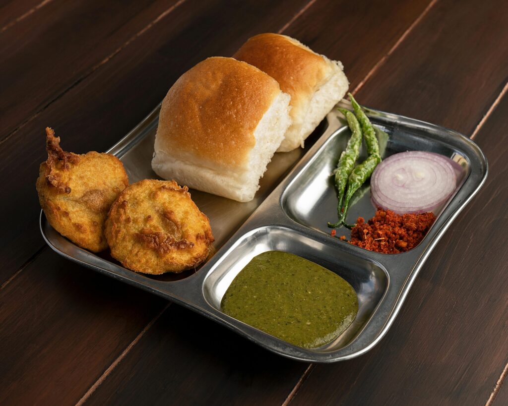 Traditional Indian vada pav served with green chutney, dry garlic chutney, fried chilies, and sliced onions on a stainless-steel plate